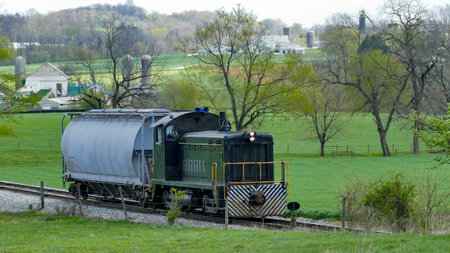 A freight train moves along the railway, surrounded by lush green fields and distant farms. The tranquil rural setting highlights the harmony between industry and nature.の写真素材