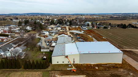 Construction workers are busy at a large warehouse site in a rural area surrounded by homes and farmland.の写真素材