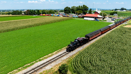 A vintage steam train moves along tracks bordered by vibrant green fields in a peaceful rural area, with a colorful train station visible in the distance under a clear blue sky.の写真素材