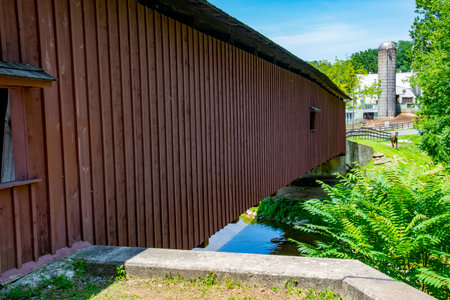 A charming wooden covered bridge extends over a crystal-clear stream, framed by vibrant foliage and a distant barn. The bright sky enhances the serene atmosphere.の写真素材