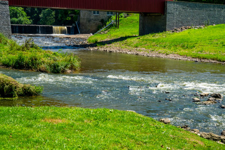 A tranquil river flows under a red covered bridge, surrounded by lush green grass and trees. Waterfalls can be seen in the background, creating a peaceful outdoor setting.の写真素材