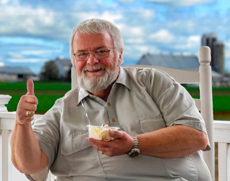 An older man with a beard sits on a porch chair, holding a bowl of dessert and giving a thumbs up. The green farmland and cloudy sky provide a peaceful backdrop.の写真素材