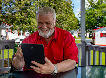 A senior man with glasses and a beard sits at a glass table in a garden, smiling while looking at a tablet. The bright sun highlights the pleasant outdoor environment.の写真素材