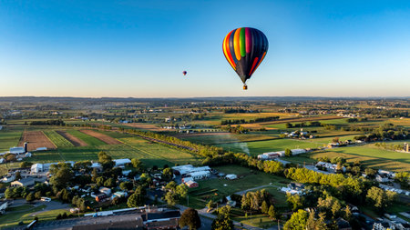 Two vibrant hot air balloons ascend gracefully over the serene countryside, capturing the beauty of expansive fields and farms under the clear blue sky during early morning light.の写真素材