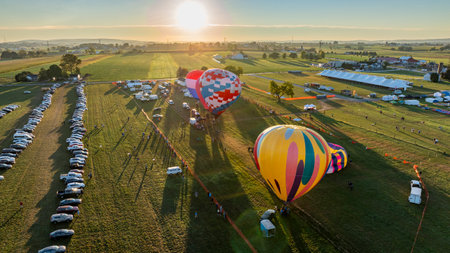Hot air balloons in vibrant colors are being inflated in a vast field during early morning. Vehicles are parked nearby as sunlight bathes the landscape in warm tones.の写真素材