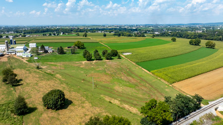 Vast farmland stretches across the scene, displaying lush green fields, crops, and trees under a bright blue sky. Nearby, farming buildings indicate agricultural activity.の写真素材