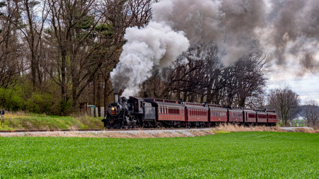 A vintage steam train plows through vibrant green fields under a clear blue sky. The engine billows white smoke as it moves along the track in the countryside during spring.の写真素材