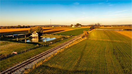A vintage steam train moves along the track, surrounded by vibrant fields and a tranquil pond. The warm light of sunset casts a golden hue over the expansive countryside.の写真素材