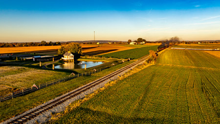 A train moves along a railroad track through expansive fields, reflecting the warm glow of the setting sun. A serene pond and quaint structures enhance the picturesque landscape.の写真素材