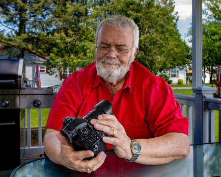 An older man with a beard sits on a porch, carefully inspecting his camera. The sun shines down on a well-kept yard in a peaceful suburban setting, evoking a sense of leisure.の写真素材