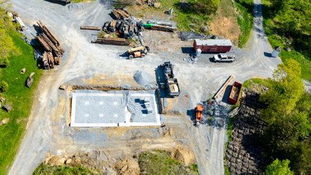 A construction site shows a freshly laid concrete foundation surrounded by piles of building materials. Heavy machinery and vehicles are present, indicating active development in a rural setting.の写真素材
