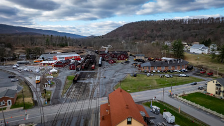 A view of a historic railway station surrounded by vintage train cars and buildings nestled in a valley. The location is peaceful, with mountains in the background under a partly cloudy sky.の写真素材