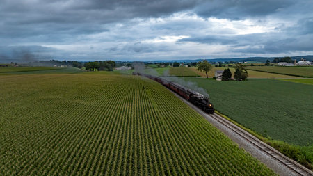 A vintage steam locomotive chugs along a winding track surrounded by vibrant green fields in Pennsylvania. Dark clouds loom overhead, creating a dramatic sky.の写真素材