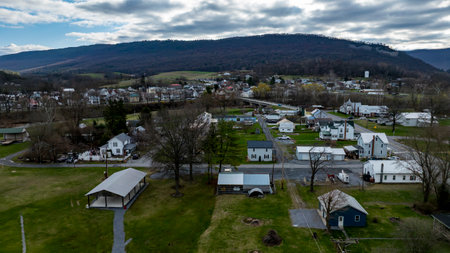 Aerial view showcases a peaceful rural town nestled among rolling hills. The landscape is dotted with houses and fields, and the sky is overcast, adding a serene touch to the scenery.の写真素材
