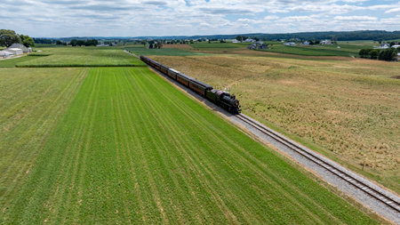 A steam train glides along the railway, surrounded by expansive green fields and distant hills under a partly cloudy sky. This serene countryside captures the beauty of rural life.の写真素材