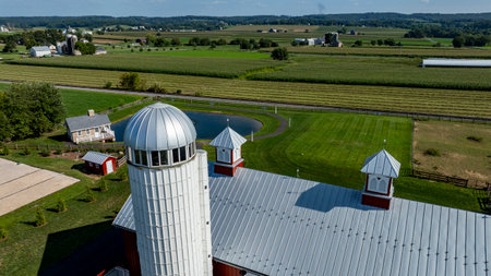 A red barn with a silver roof stands tall, surrounded by vibrant green fields and a small pond. Nearby, the countryside stretches into the horizon, showcasing peaceful rural scenery.の写真素材