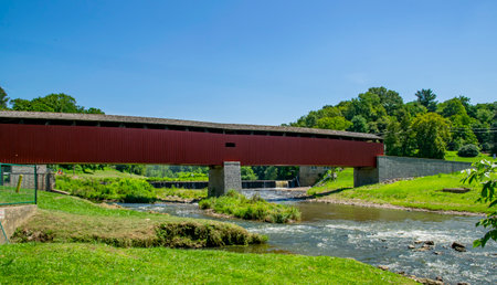 A red covered bridge crosses over a gentle stream surrounded by vibrant green trees on a bright, sunny day. Nature thrives in this peaceful rural setting, inviting exploration.の写真素材