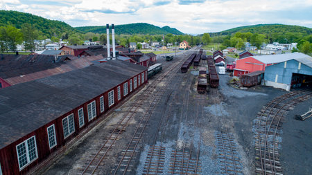 A vintage railway yard features numerous freight cars and wooden warehouses under a bright sky. Lush green hills surround the area, creating a picturesque rural setting.の写真素材