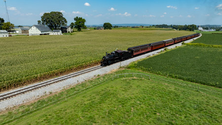 A vintage steam train navigates through lush green fields in Pennsylvania, surrounded by farmland. The clear blue sky above adds to the charm of this picturesque rural setting.の写真素材
