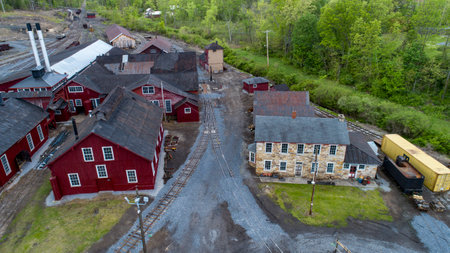 A former industrial complex with red and stone buildings is set amidst vibrant green trees in the daytime. Train tracks wind through the grounds, showcasing the site's historic charm and landscape.の写真素材
