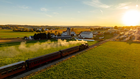 A vintage steam train moves along tracks amidst green fields and farmland. The sun sets, casting warm light on the rural landscape, highlighting barns and silos.の写真素材