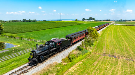 Vintage Steam Train Travels Through Lush Green Fields in Rural Landscape During Daytime. High quality photoA vintage steam train moves along a scenic track surrounded by vibrant green fields. Bright skies enhance the picturesque rural landscape, creating a nostalgic atmosphere.の写真素材