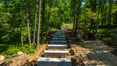 A construction site shows workers laying concrete slabs to form a walking path through a lush wooded area. Heavy machinery is present, and trees surround the site, enhancing the natural landscape.の写真素材