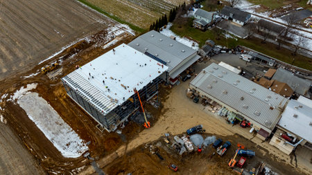 Workers are busy on the roof of a large commercial building, while heavy machinery and materials are scattered around the site. Snow covers parts of the ground in a rural landscape.の写真素材