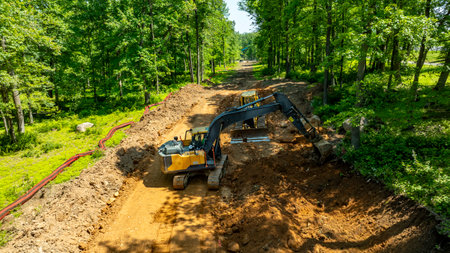Excavators work on clearing land in a densely wooded area. The operators maneuver heavy equipment to prepare the site for future development under clear blue skies.の写真素材