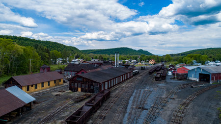 An expansive railway yard features vintage buildings and long tracks surrounded by lush trees and hills. The scene captures the remnants of a bustling transportation hub under a dramatic sky.の写真素材