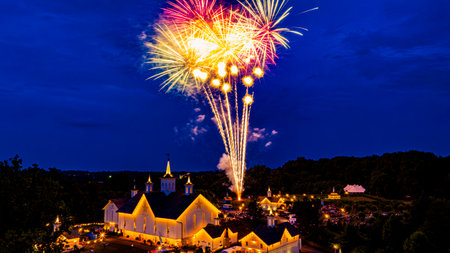 Bright fireworks light up the night sky above a white church surrounded by trees, as people gather below, enjoying a summer celebration filled with joy and excitement.の写真素材