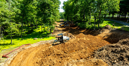 Heavy machinery operates in a forested area, moving dirt and reshaping the landscape. Workers are seen in the background, contributing to the construction project during daylight.の写真素材