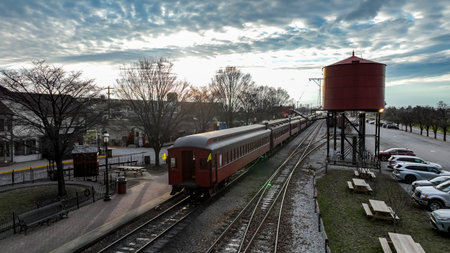 A vintage train moves through a serene town as the sun sets, casting a warm glow over the landscape, with a water tower and parked cars in the foreground.の写真素材