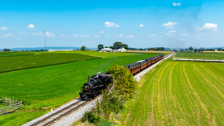 A steam locomotive pulls a long train through expansive green fields and lush farmland. The bright...