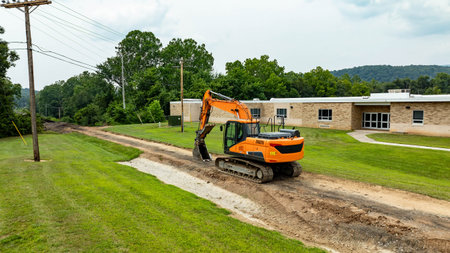 A large excavator works on a dirt road near a school building. The area is green with trees in the...