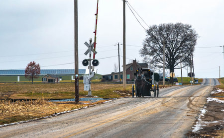 A horse-drawn buggy moves along a dirt road near a train crossing. Power lines line the way as the buggy approaches a farmhouse. It is a cloudy winter day with few people around.の写真素材