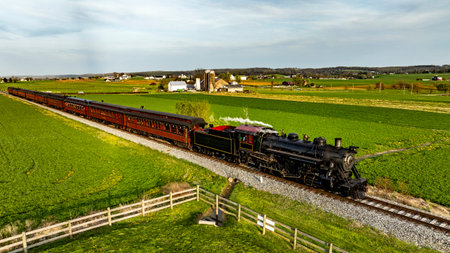 A steam train moves along tracks in a rural area. The train runs through green fields with houses in...