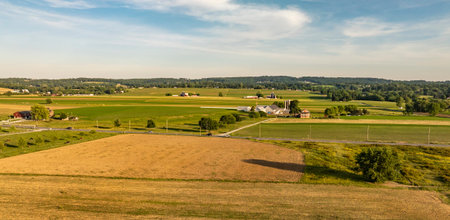 A wide view shows fields and small houses spread across a rural landscape. Green fields and patches of brown soil are visible under a blue sky with white clouds.