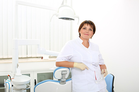 female dentist holding safety glasses and standing in the dental office leaning on the dental chairの写真素材