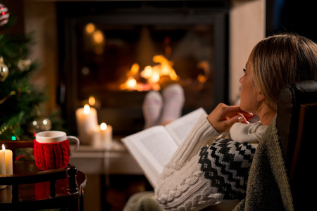 Woman is resting with cup of hot drink and book near the fireplaceの写真素材
