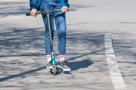The legs of an unknown girl in white sneakers and tight jeans ride on a black electric scooter over...