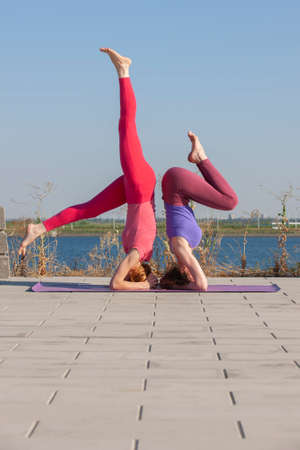 Two Caucasian Females in tights and sneakers Doing Yoga Exercises on the background of the river. Practicing and creating a showy composition while standing in the pose of an athlete's bridge.の写真素材
