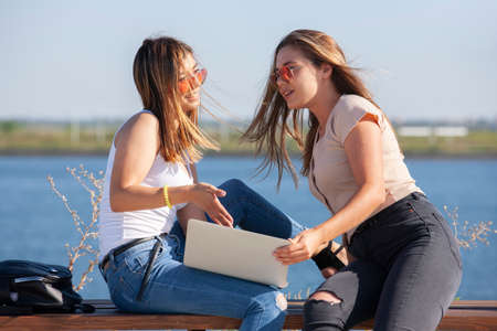 Two beautiful young caucasian girls looking amazed to a laptop while sitting outside in the park on a bench. Portrait.の写真素材