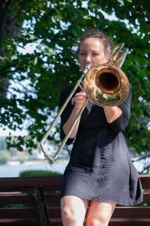 Girl learning to play trombone. Girl plays leaning on a park bench. Portrait.の写真素材