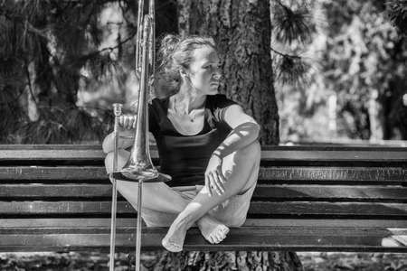 Girl learning to play trombone. Girl plays sitting on a park bench. Portrait.の写真素材