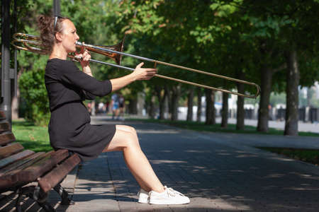 Girl learning to play trombone. Girl plays sitting on a park bench. Portrait.の写真素材