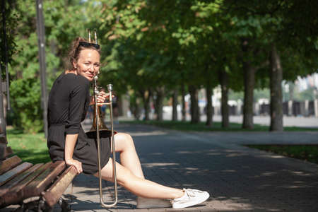 Girl learning to play trombone. Girl plays sitting on a park bench. Portrait.の写真素材