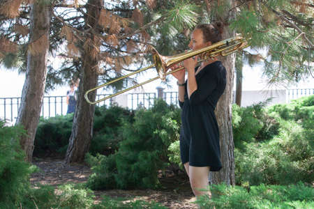 Girl learning to play trombone. Girl plays standing on the alley of a city park. Portrait.の写真素材