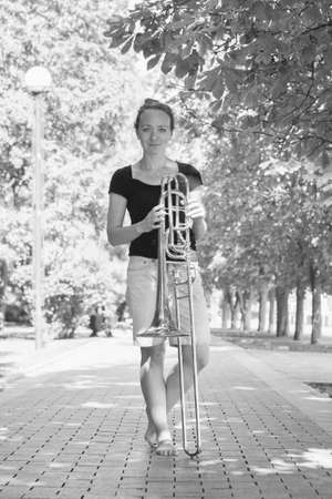 Girl learning to play trombone. Girl plays standing on the alley of a city park. Portrait.の写真素材