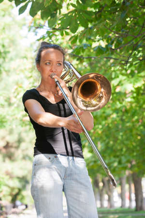 Girl learning to play trombone. Girl plays standing on the alley of a city park. Portrait.
